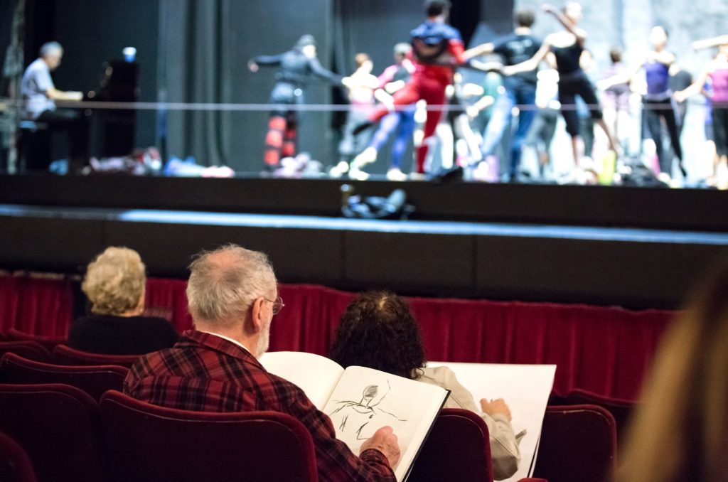 3 people aged over 70 years old sat in theatre seats, whilst around 12 dancers rehearse on stage. The people sat in their seats have large sketch-pads and are drawing what they see
