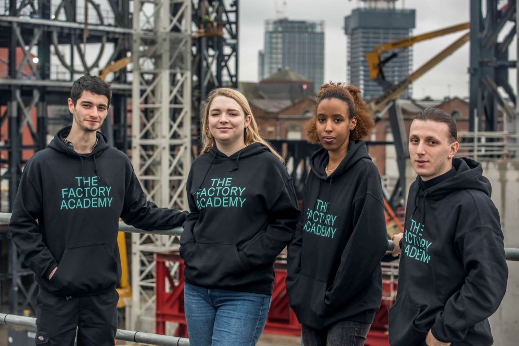 Four young people wearing jumpers emblazoned with the words 'The Factory Academy' stood with The Factory construction site in the background 