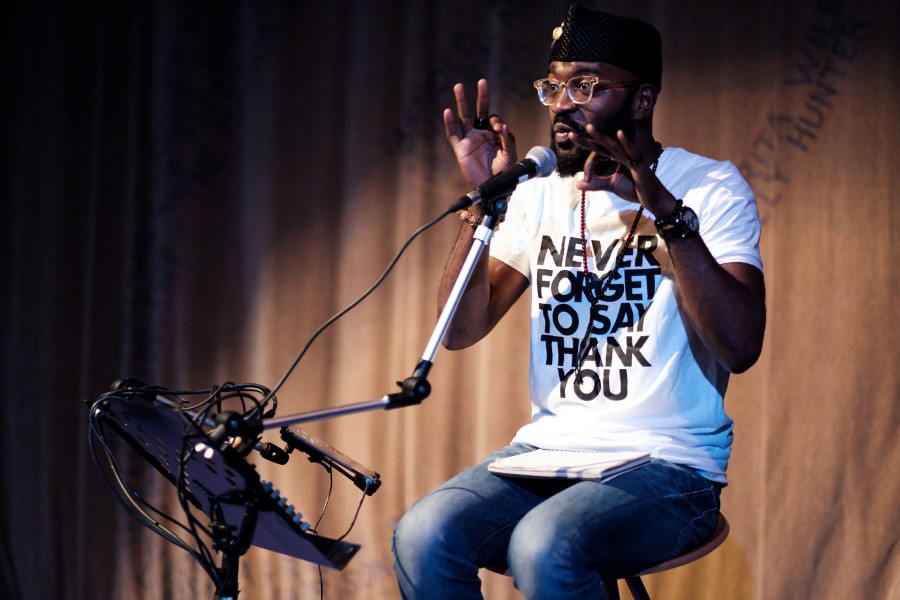 Inua Ellams sat on a stool. He's talking into a microphone, has his hands raised almost to his face making an 'OK' gesture with both hands. He's wearing a white t-shirt with 'NEVER FORGET TO SAY THANK YOU' emblazoned across it. 